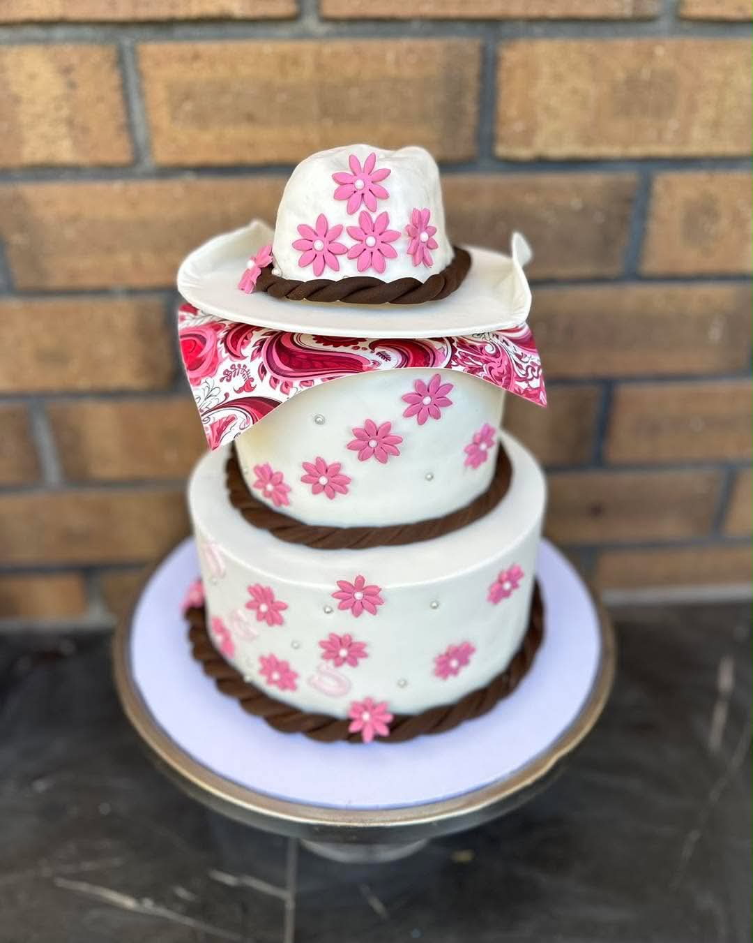 Two-tier white fondant cowgirl birthday cake with fondant cowboy hat topper, pink daisy flowers, brown rope borders and paisley bandana wrap